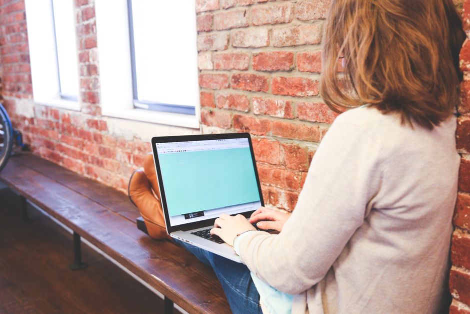 A woman is using a laptop for remote work while sitting indoors against a brick wall.